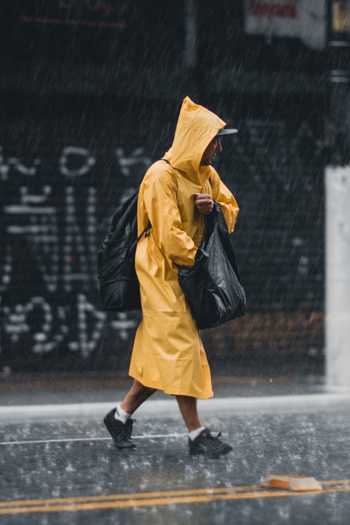 A man walks through city streets in pouring rain, clad in a yellow raincoat, carrying bags.